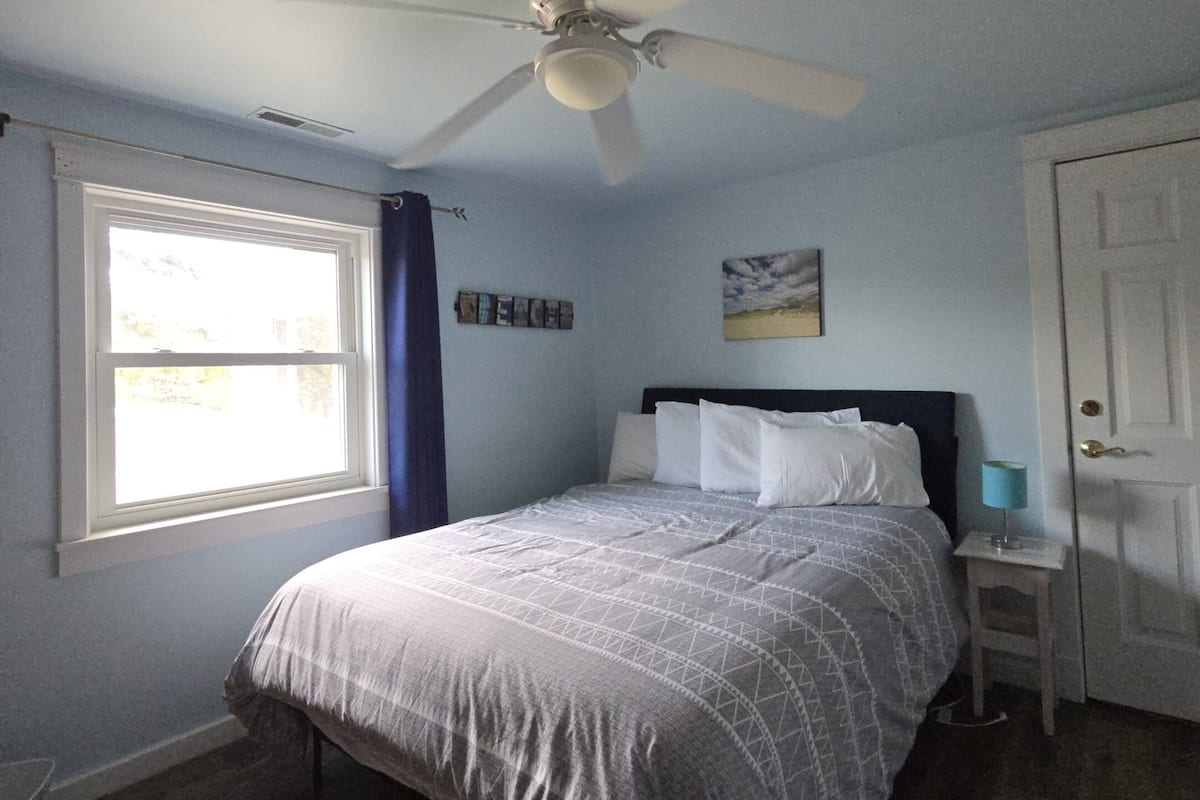 Une chambre à coucher comprenant un lit, un ventilateur de plafond, une fenêtre avec des rideaux, une porte et une petite table de chevet avec une lampe.