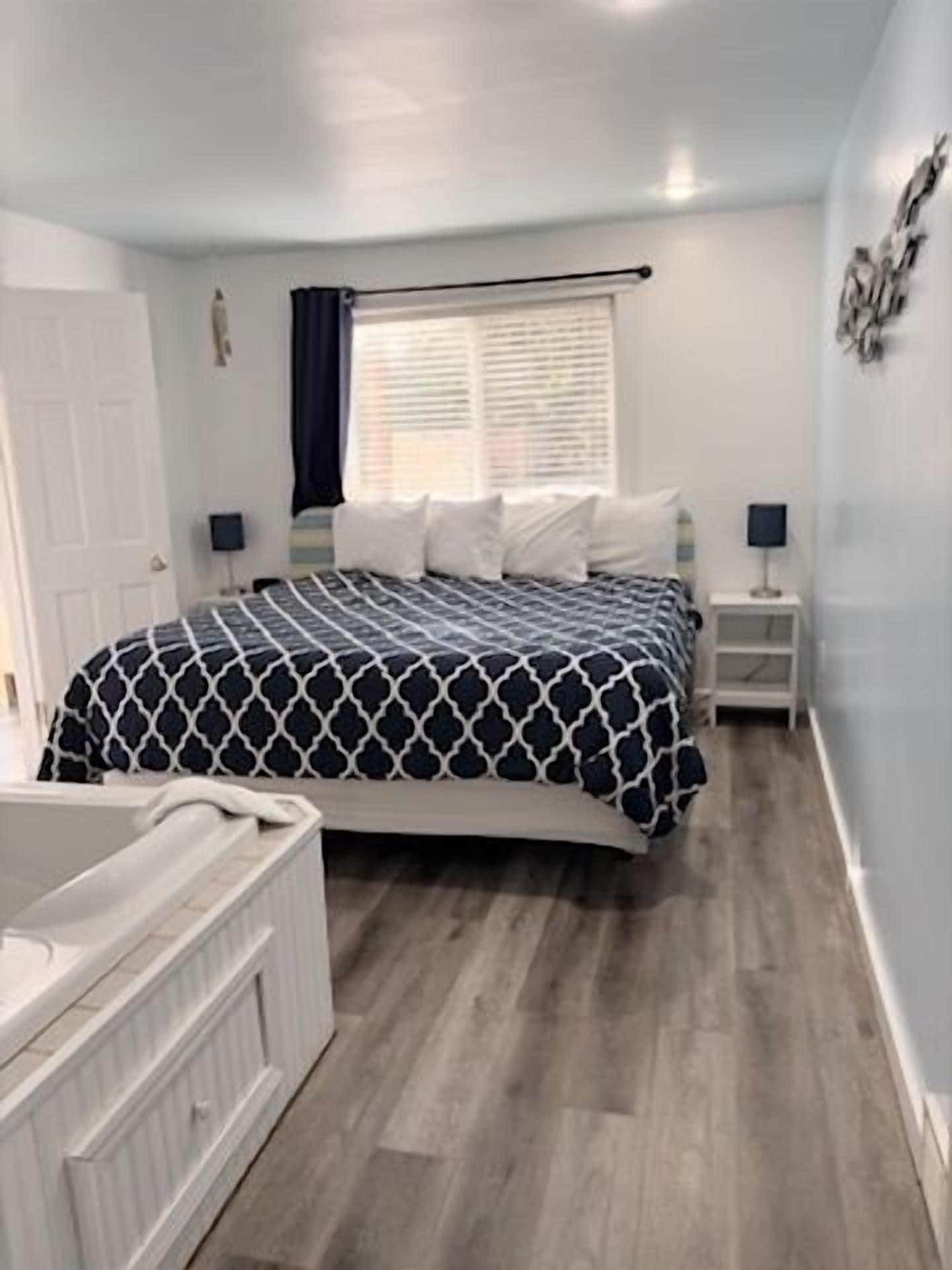 Um quarto com cama, mesa de cabeceira, janela com persianas, porta e cômoda branca.