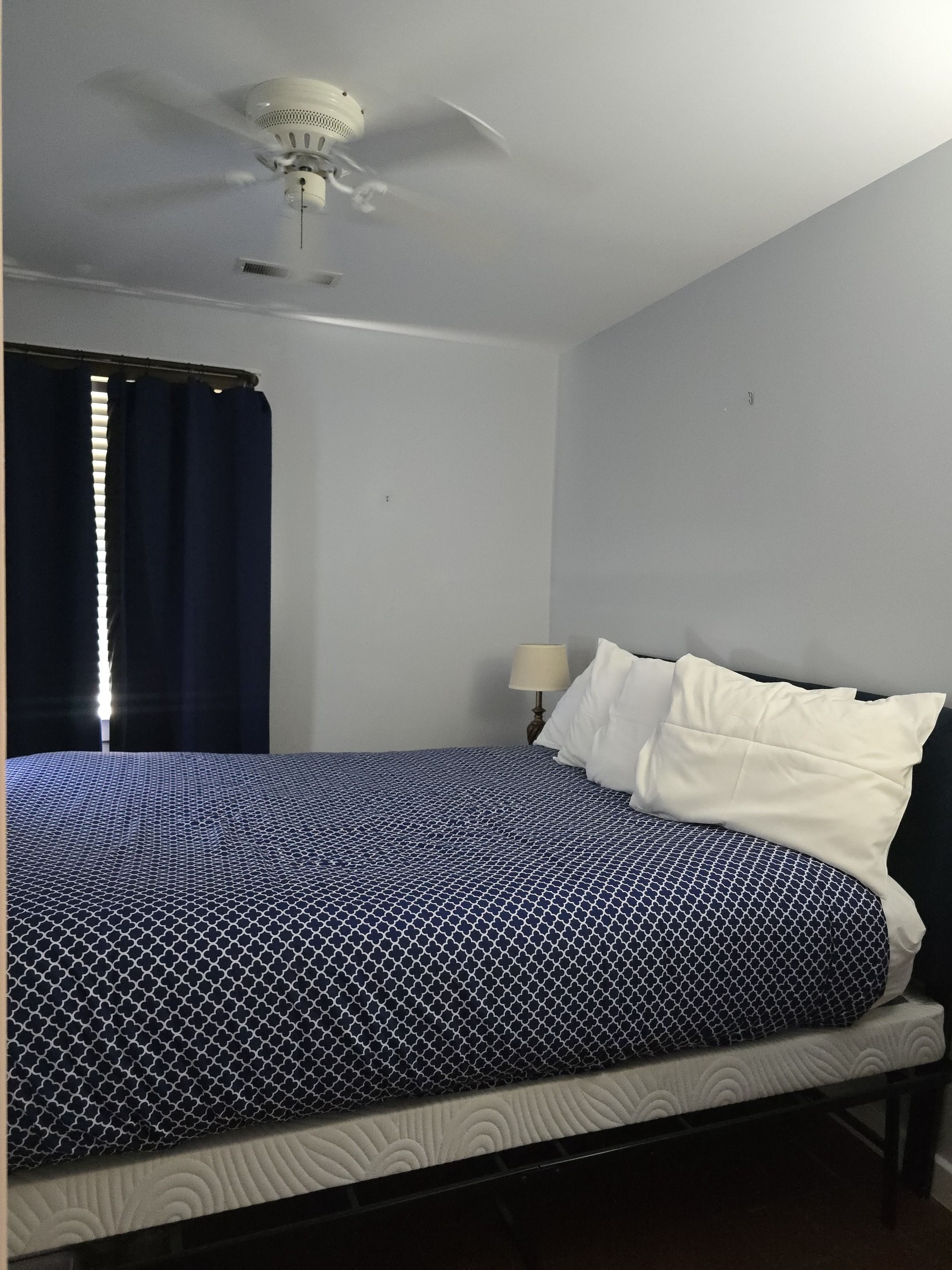 Una habitación con una cama, un ventilador de techo, una ventana con cortinas y una lámpara de noche.