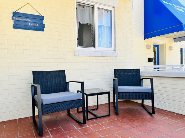 Deux fauteuils en osier avec des coussins, un mur en briques blanches et un auvent bleu.