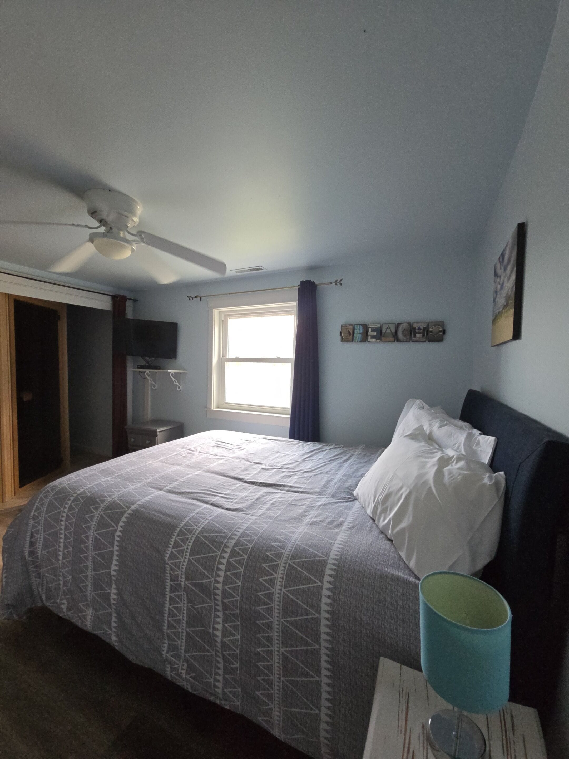 A bedroom with a bed, a ceiling fan, a window with curtains, a wall-mounted TV, and a small table with a lamp.