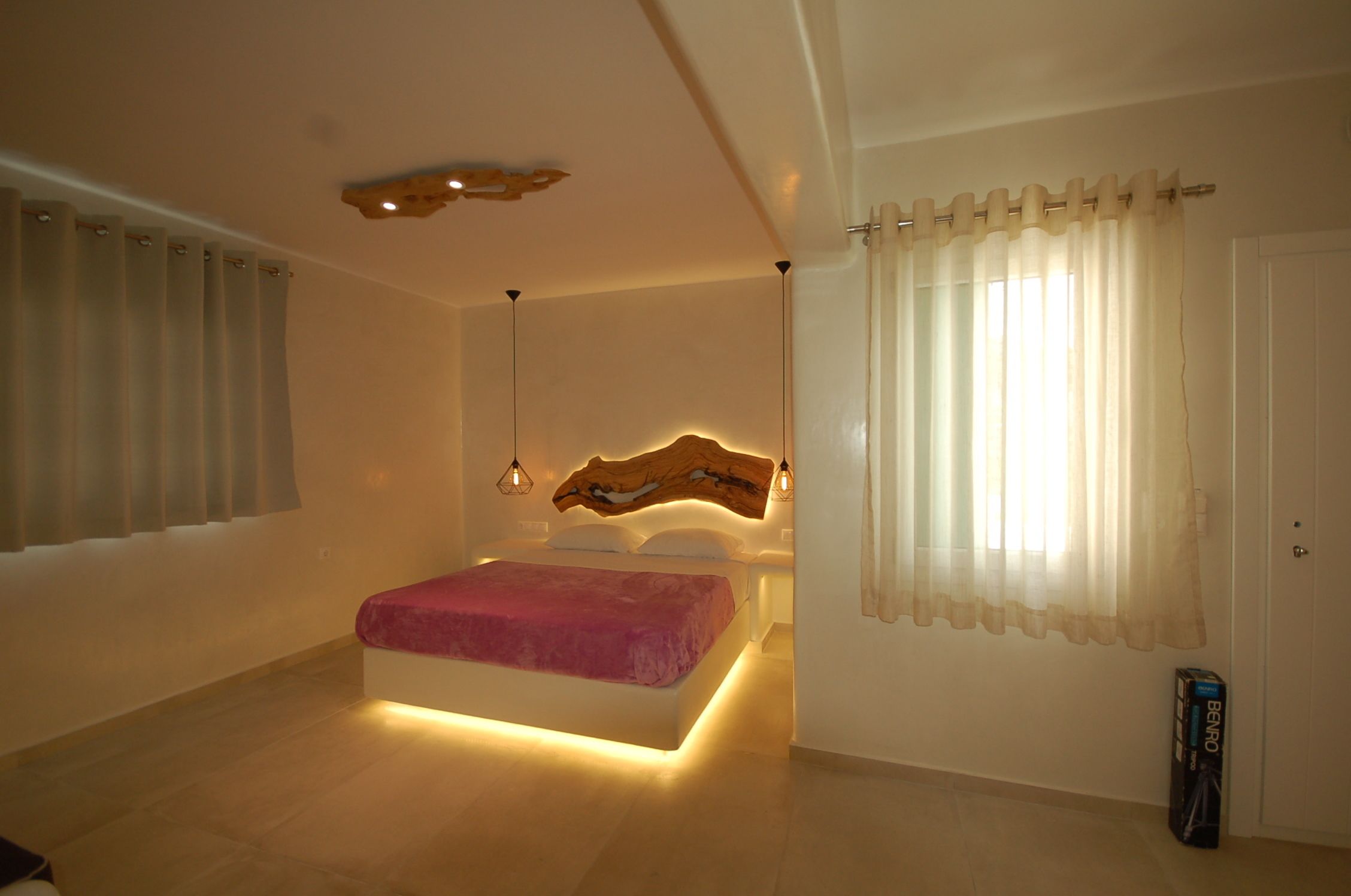 A bedroom with a bed, a wooden headboard, a bedside table, a nightstand, a window with sheer curtains, and a door.