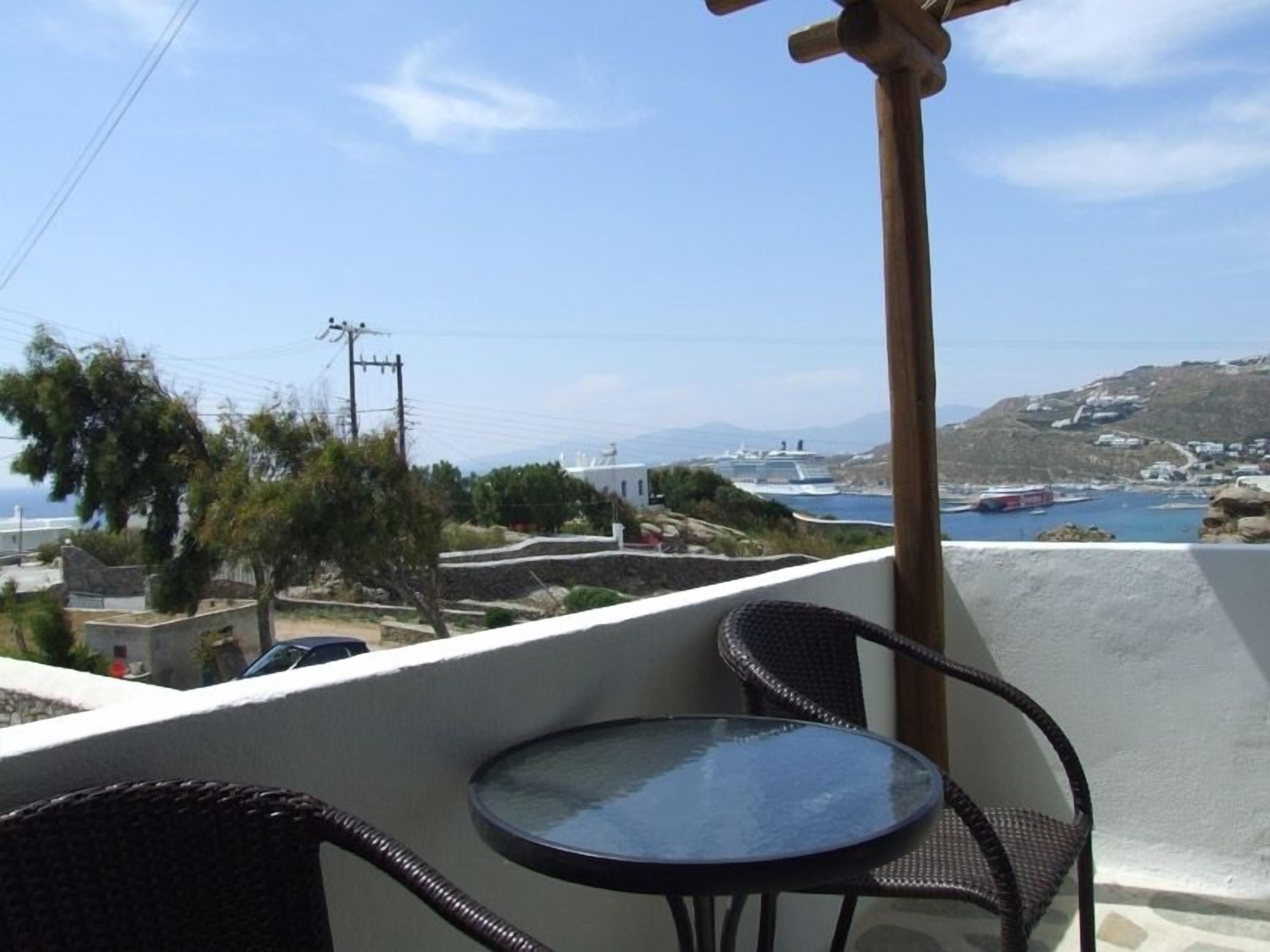 A balcony with a round glass table and two wicker chairs, overlooking a coastal area with boats and buildings.