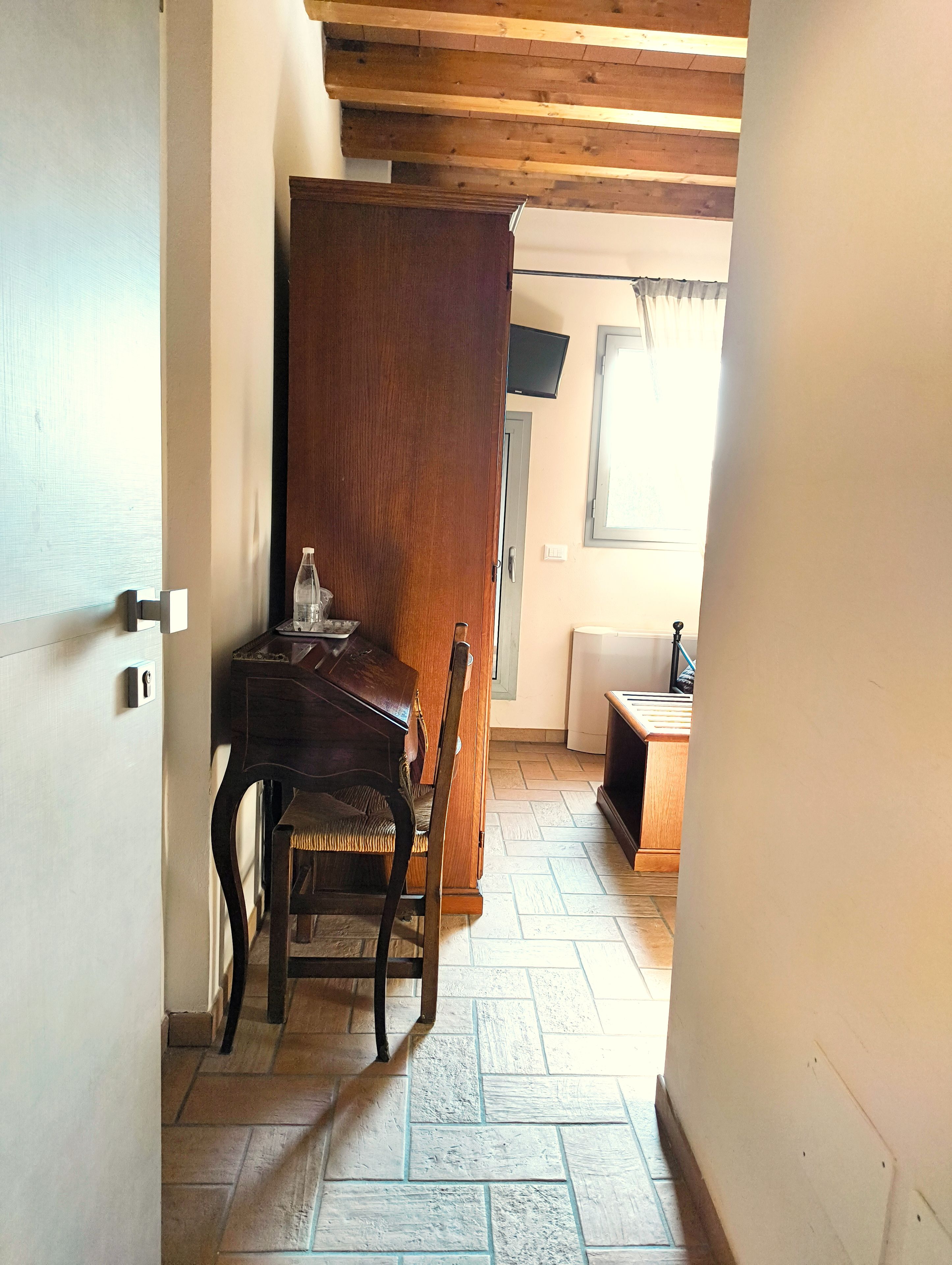 A room with a wooden door, a small table with a chair, a bottle, and a television on the wall.