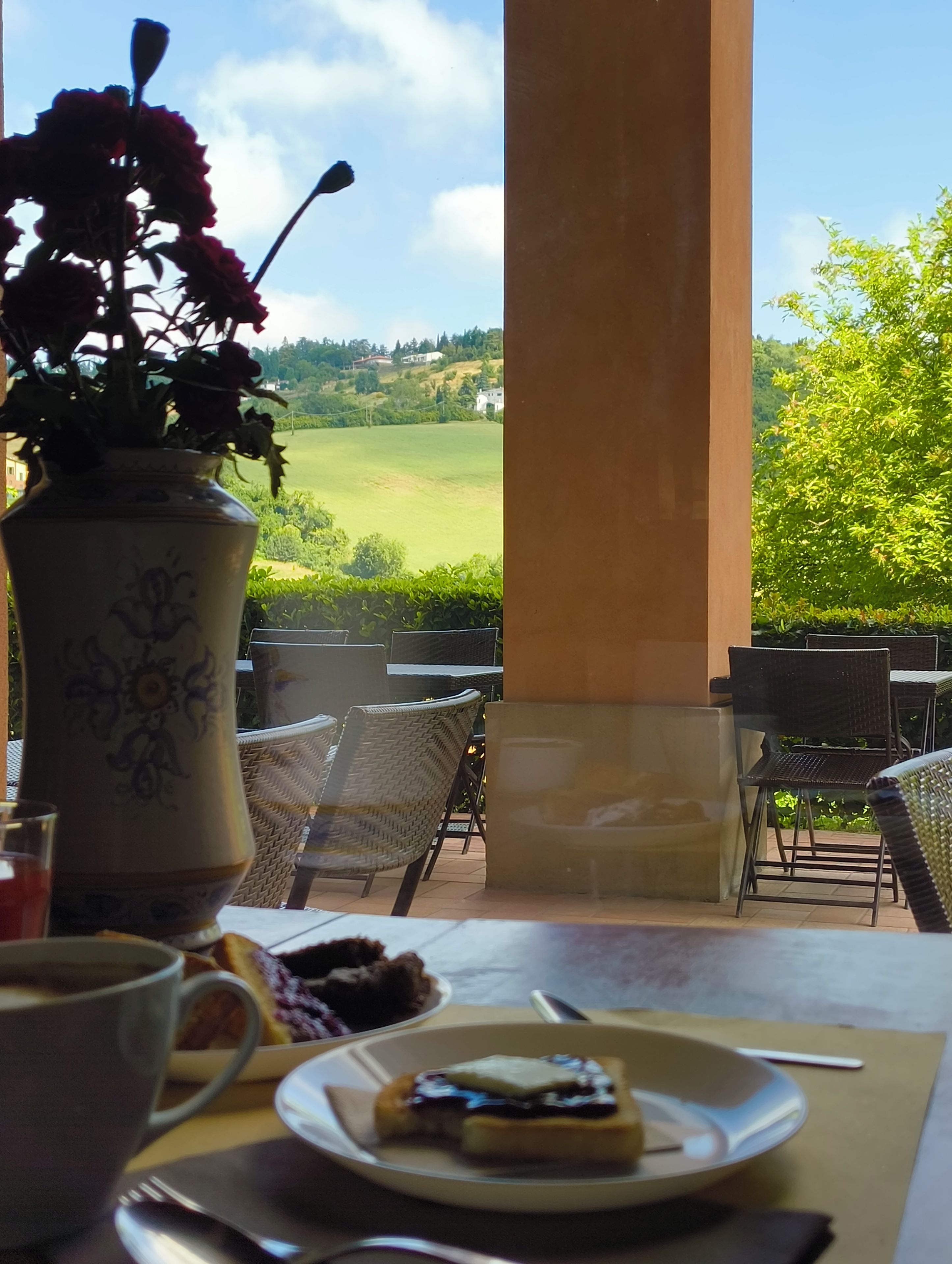 An outdoor dining area with a table, chairs, a vase with flowers, and a view of a green landscape.