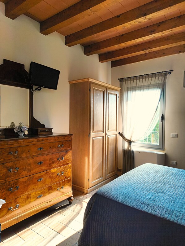 Un dormitorio con un aparador de madera, una cama con colcha azul, un armario de madera, un televisor montado en la pared y una ventana con cortinas.