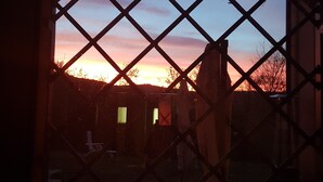Uitzicht door een raam met een ruitpatroon, waarop een zonsondergang te zien is met een gradiënte lucht en silhouetten van bomen en gebouwen.