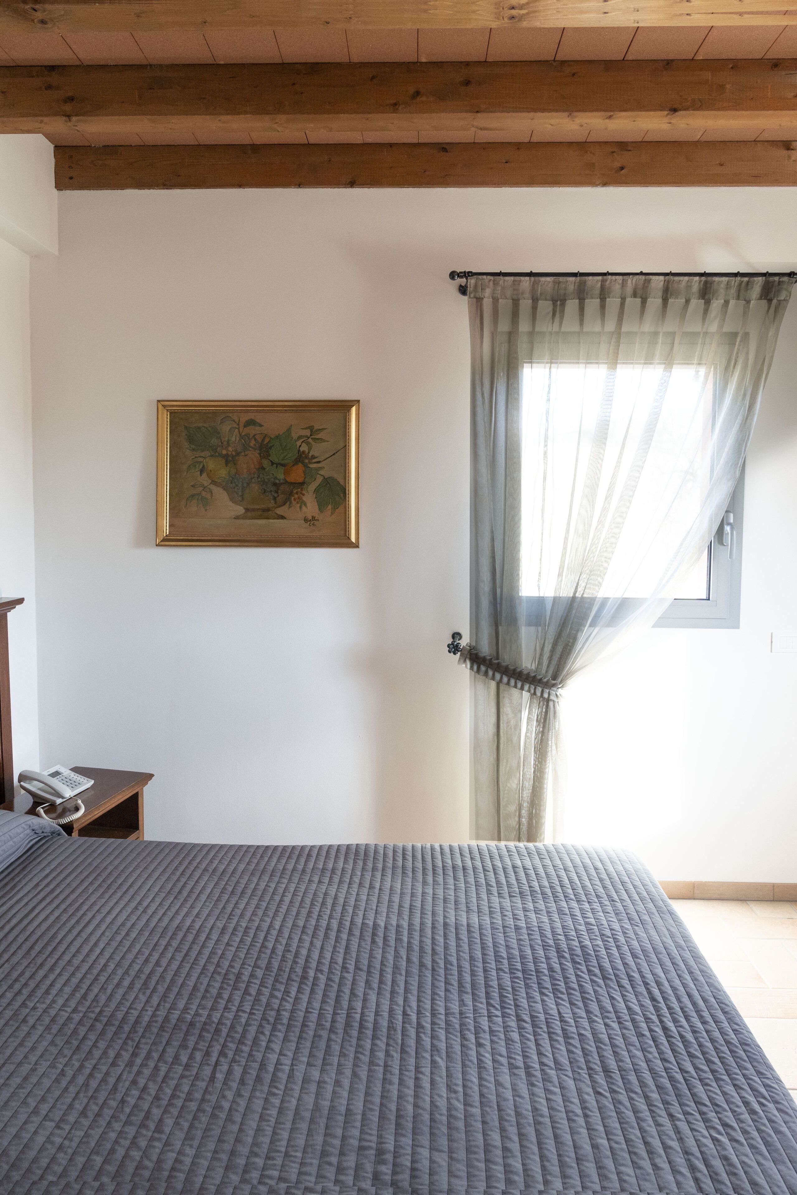 A bedroom with a bed, a bedside table, a telephone, a window with sheer curtains, and a framed painting on the wall.