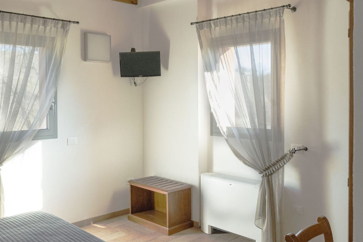 Een kamer met een bed, een houten tafel, een aan de muur bevestigde televisie en een raam met dunne gordijnen.