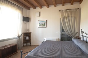A bedroom with a bed, a television, a painting, a shelf with a decorative item, and a window with curtains. - Agriturismo Ben Ti Voglio (Bologna)