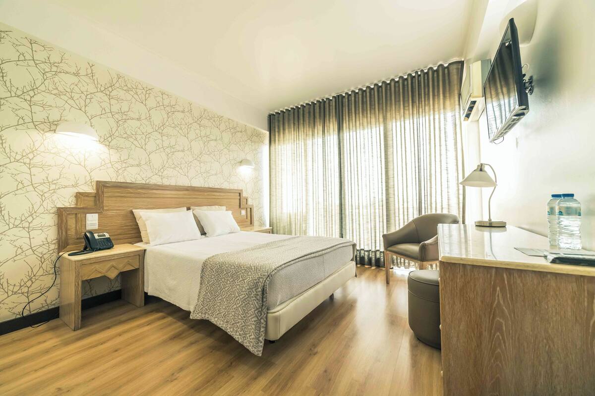 A hotel room with a bed, bedside table with a phone, a desk with a lamp, a chair, and a TV mounted on the wall.
