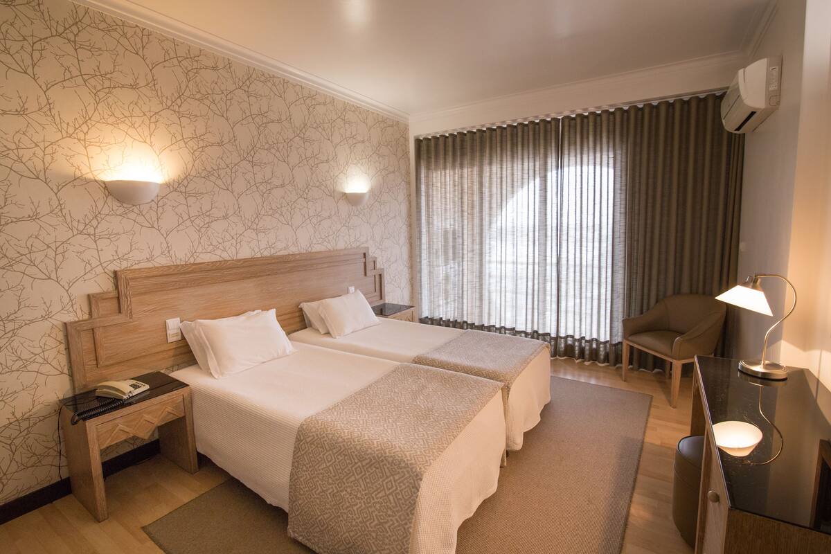 A hotel room with two beds, a desk, a chair, a lamp, and a window with curtains.