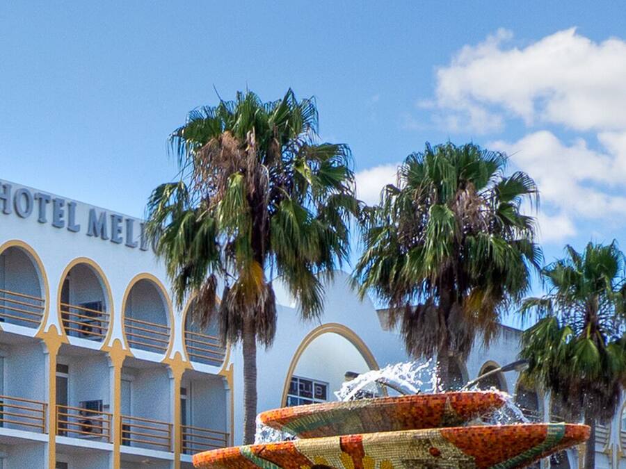 Un hotel con una fuente en primer plano, palmeras y un cielo azul despejado.