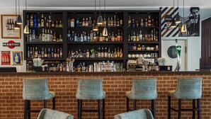Una zona de bar con pared de ladrillo, tres taburetes y estantes llenos de botellas y vasos.