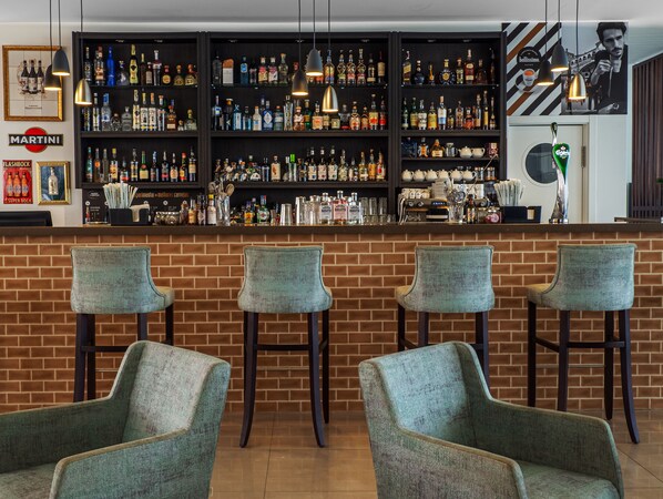 Una zona de bar con pared de ladrillo, tres taburetes y estantes llenos de botellas y vasos.
