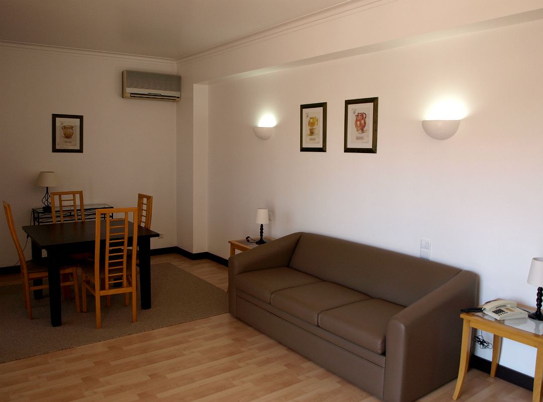 Een woonkamer met een bruine bank, een houten eettafel en stoelen. Er zijn wandlampen en afbeeldingen in lijsten.