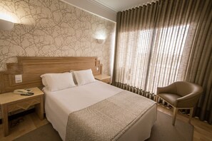 A hotel room with a large bed, a wooden headboard, a nightstand with a phone and a lamp, a chair, and a window with curtains.