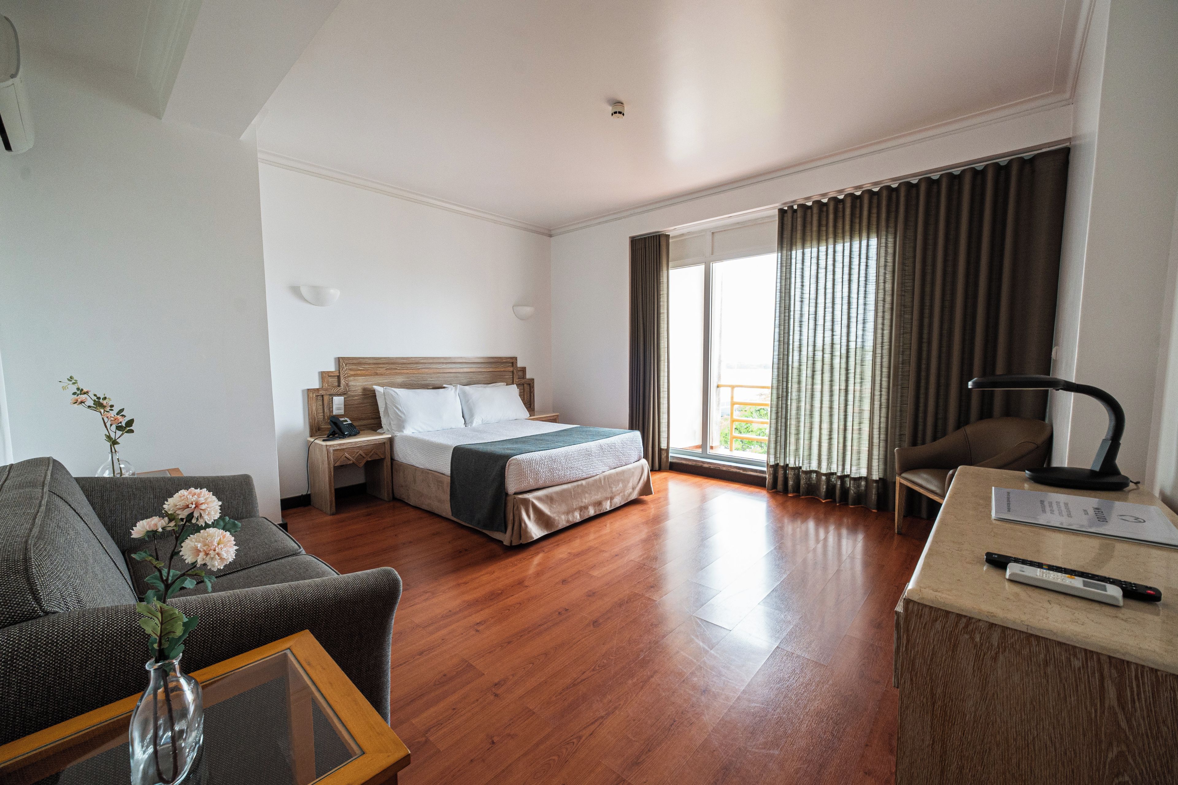 A hotel room with a bed, a desk, a chair, a vase with flowers, and a large window with curtains.