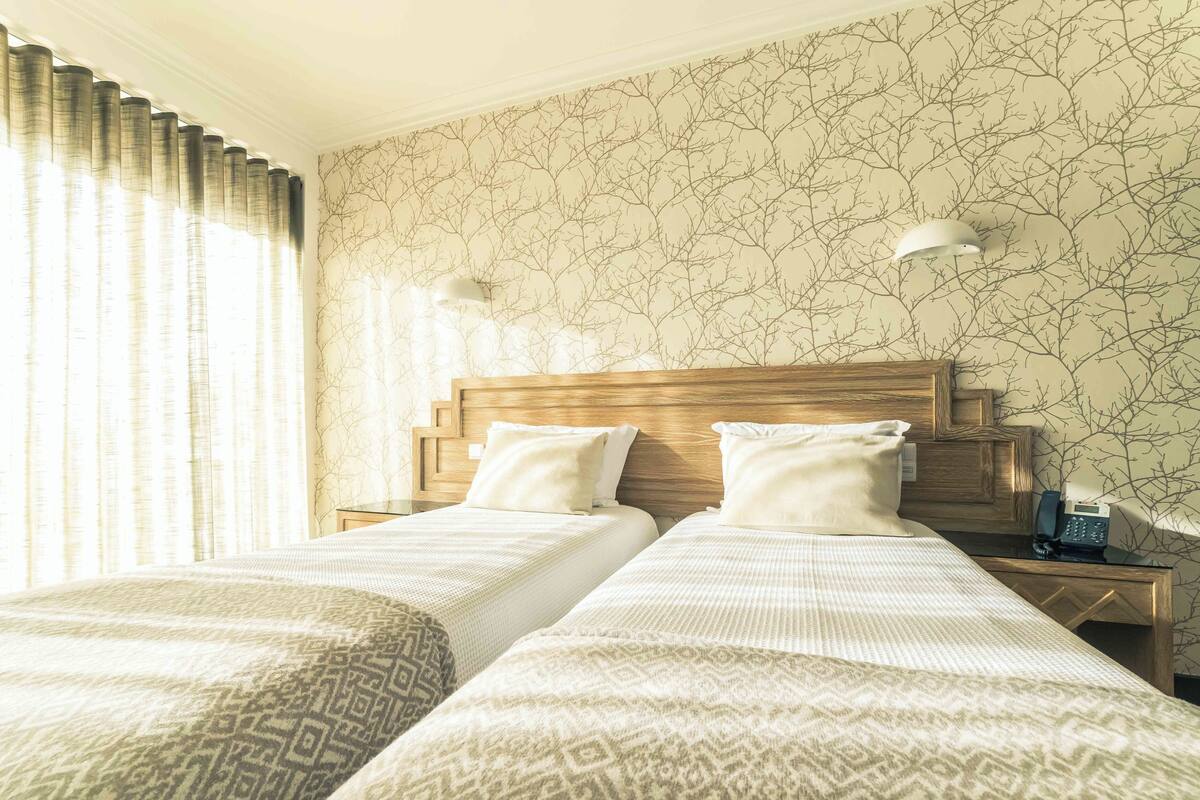 A hotel room with two beds, a wooden headboard, a telephone, and a wall with a tree branch pattern.