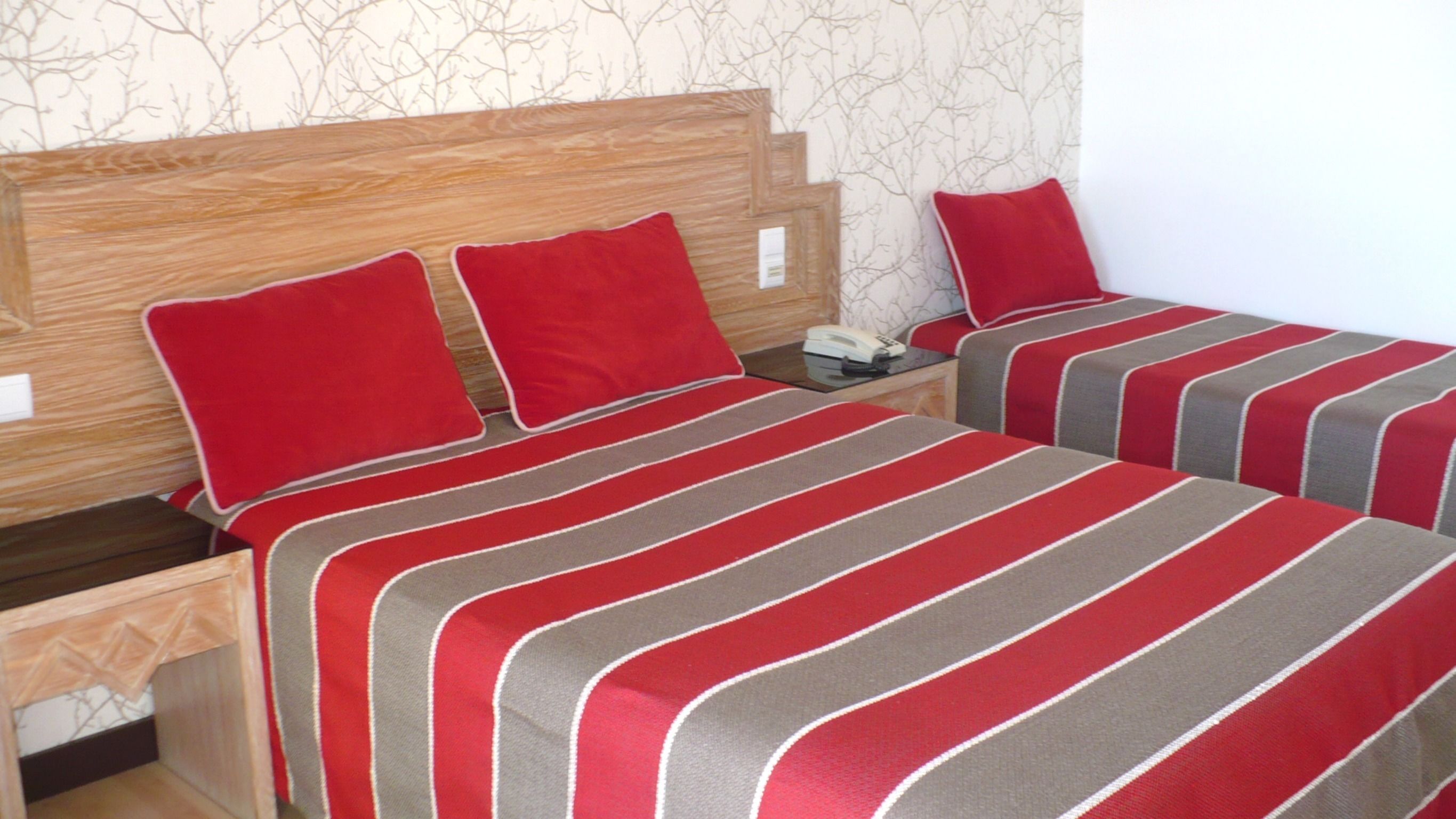 Habitación de hotel con dos camas, cada una con ropa de cama y almohadas a rayas grises y rojas. Cuenta con un cabecero de madera, una mesita de noche con un teléfono y una pequeña mesita auxiliar.