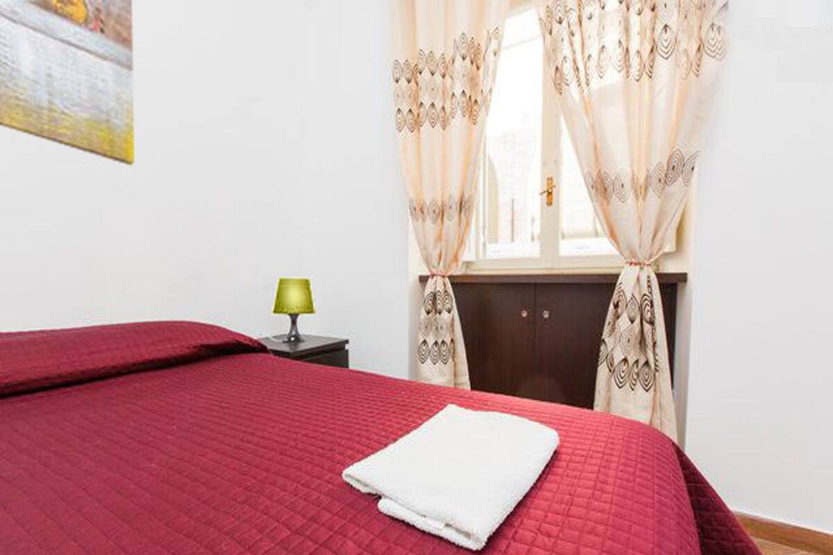A neatly made bed with a red quilt, a white towel, and a lamp on a bedside table.