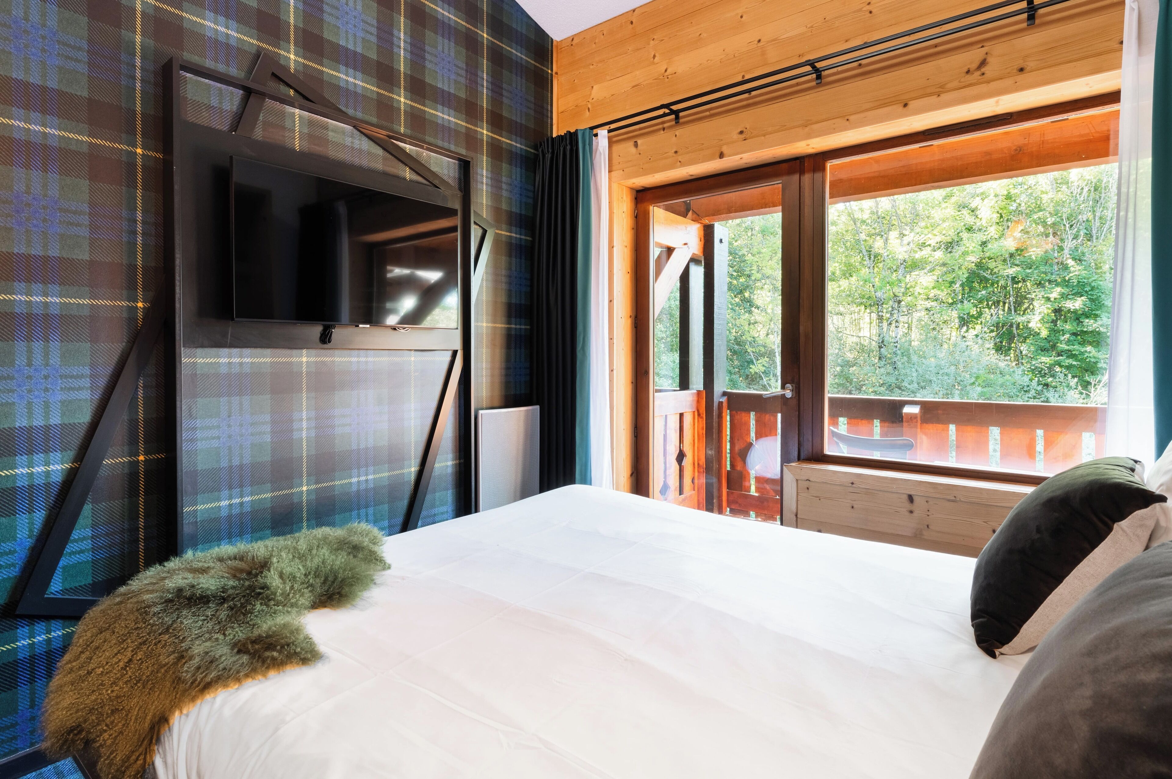 Une chambre avec un grand lit, un téléviseur fixé au mur et une fenêtre donnant sur les arbres.