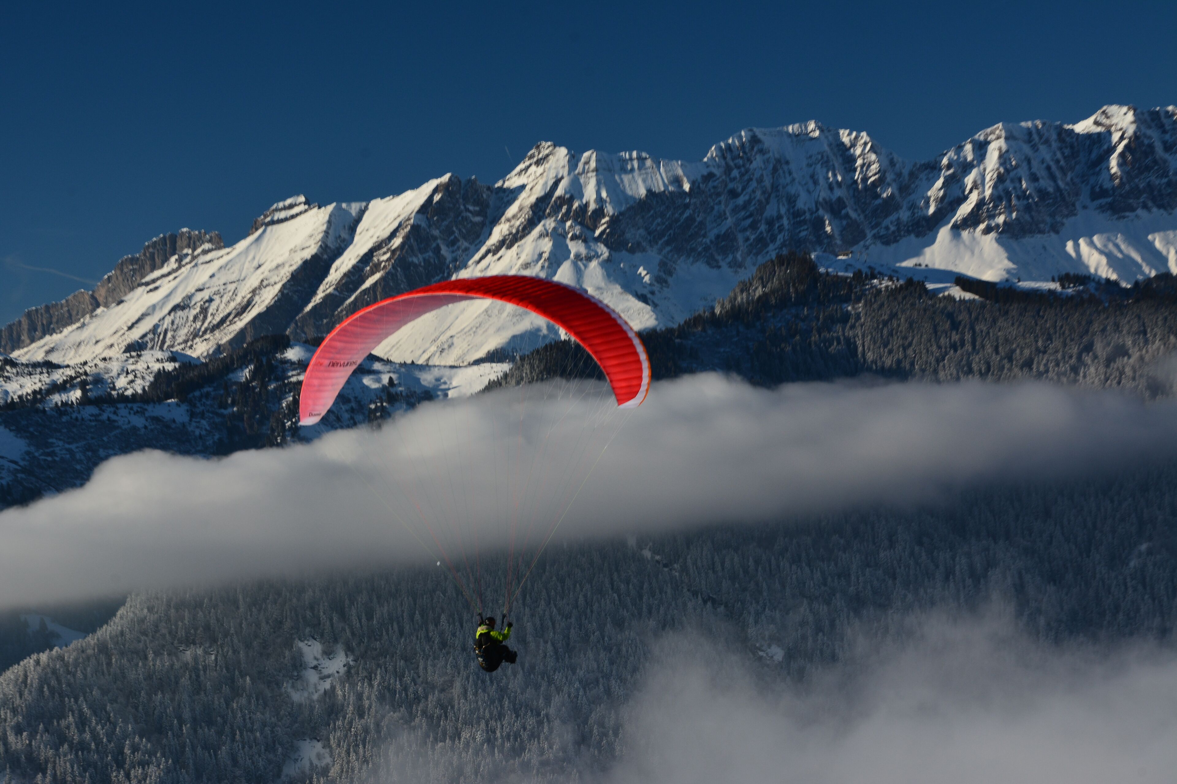 Ein Gleitschirmflieger überfliegt ein verschneites Gebirge mit einem bewaldeten Tal darunter.