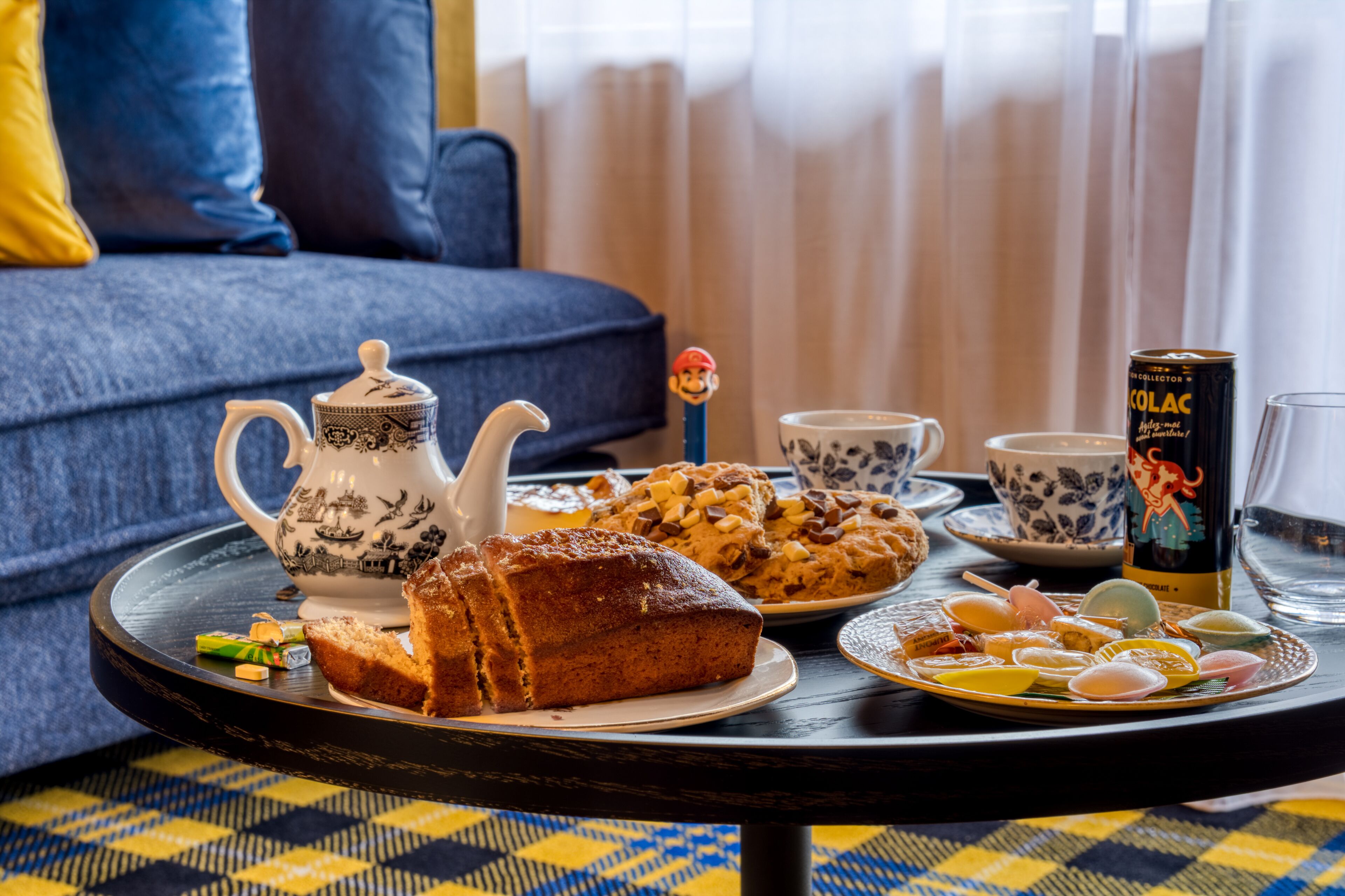 Une table à manger avec une théière, une part de gâteau, une assiette de biscuits, une canette de bière et un verre d’eau.