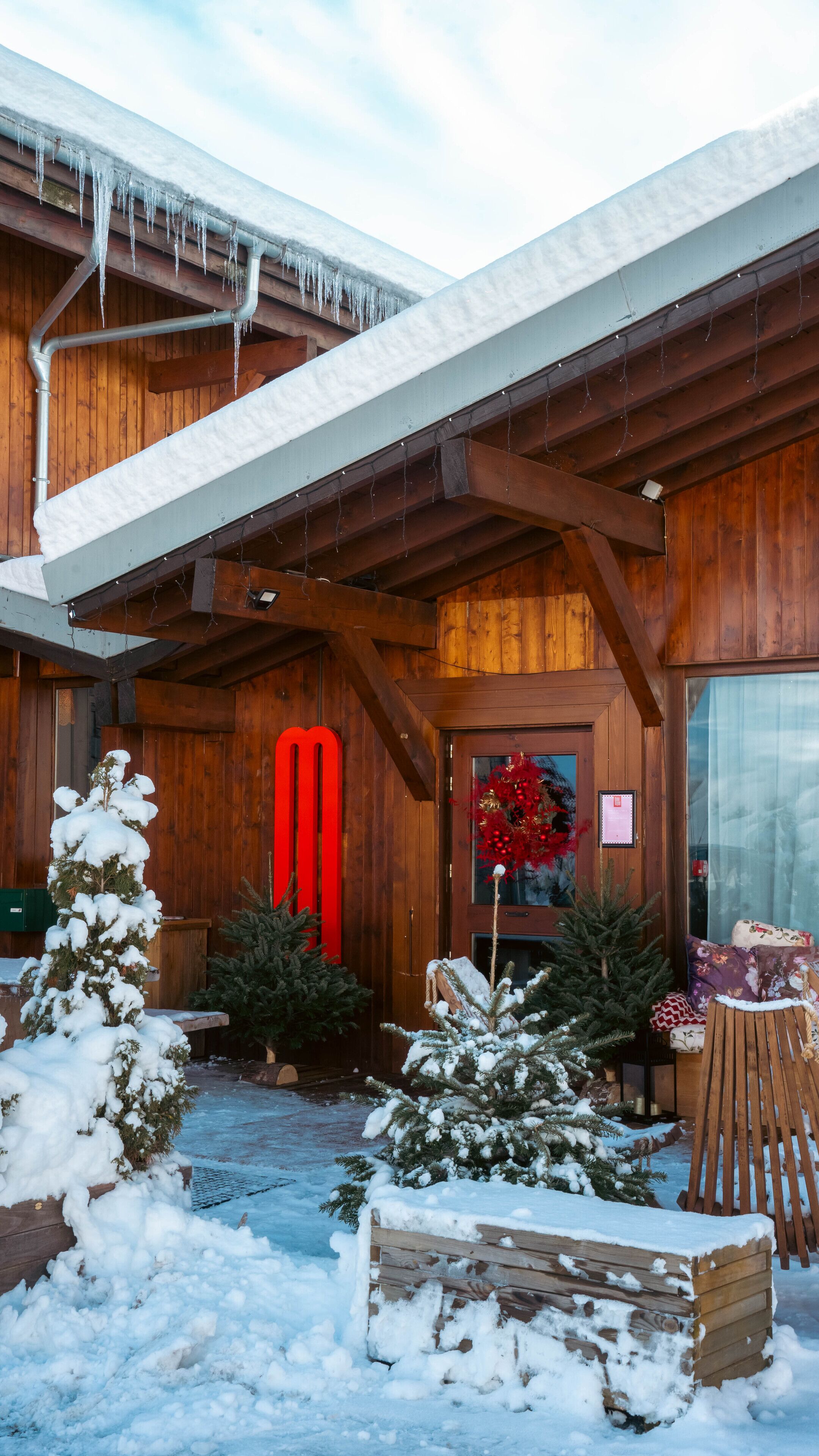 A wooden cabin with a snow-covered roof and icicles. There are Christmas trees and a bench outside.