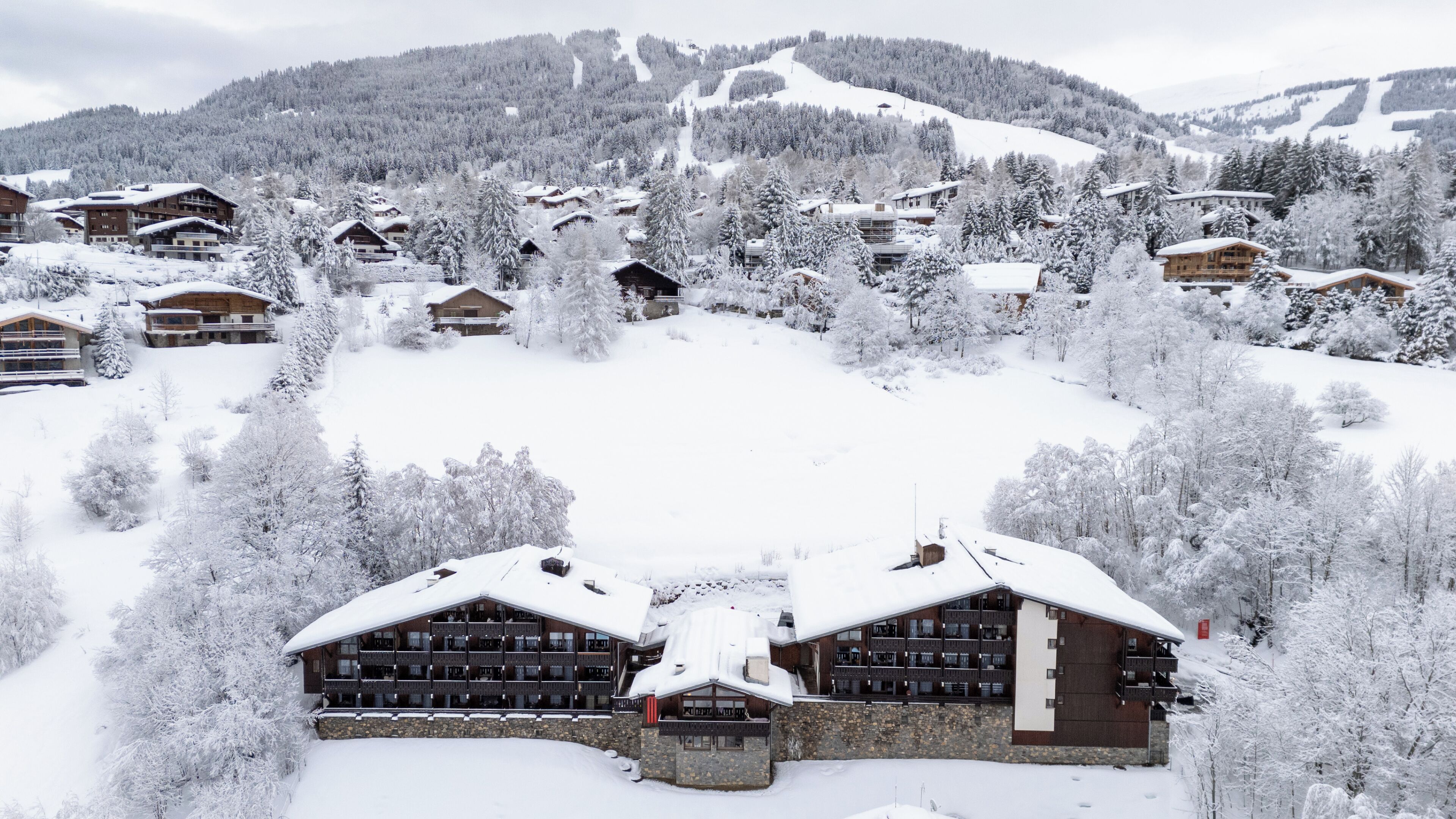 Ein mit Schnee bedecktes Chalet mit mehreren Zimmern, ein Skipisten im Hintergrund und eine gebirgige Landschaft.
