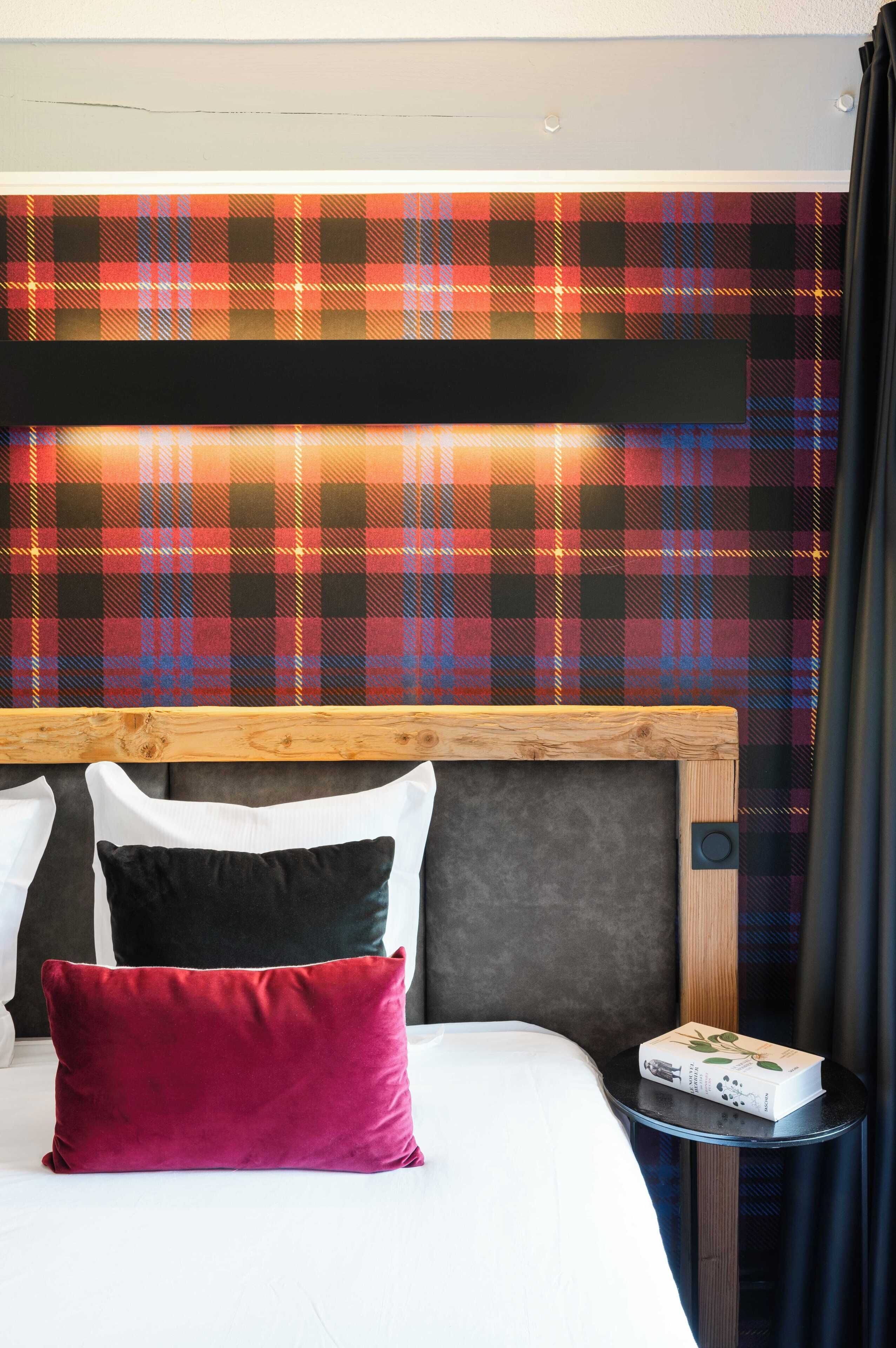 Un lit avec un oreiller rouge, un oreiller blanc et un oreiller noir. Il y a une petite table sur laquelle se trouve un livre. Le mur derrière le lit est décoré d’un motif à carreaux.