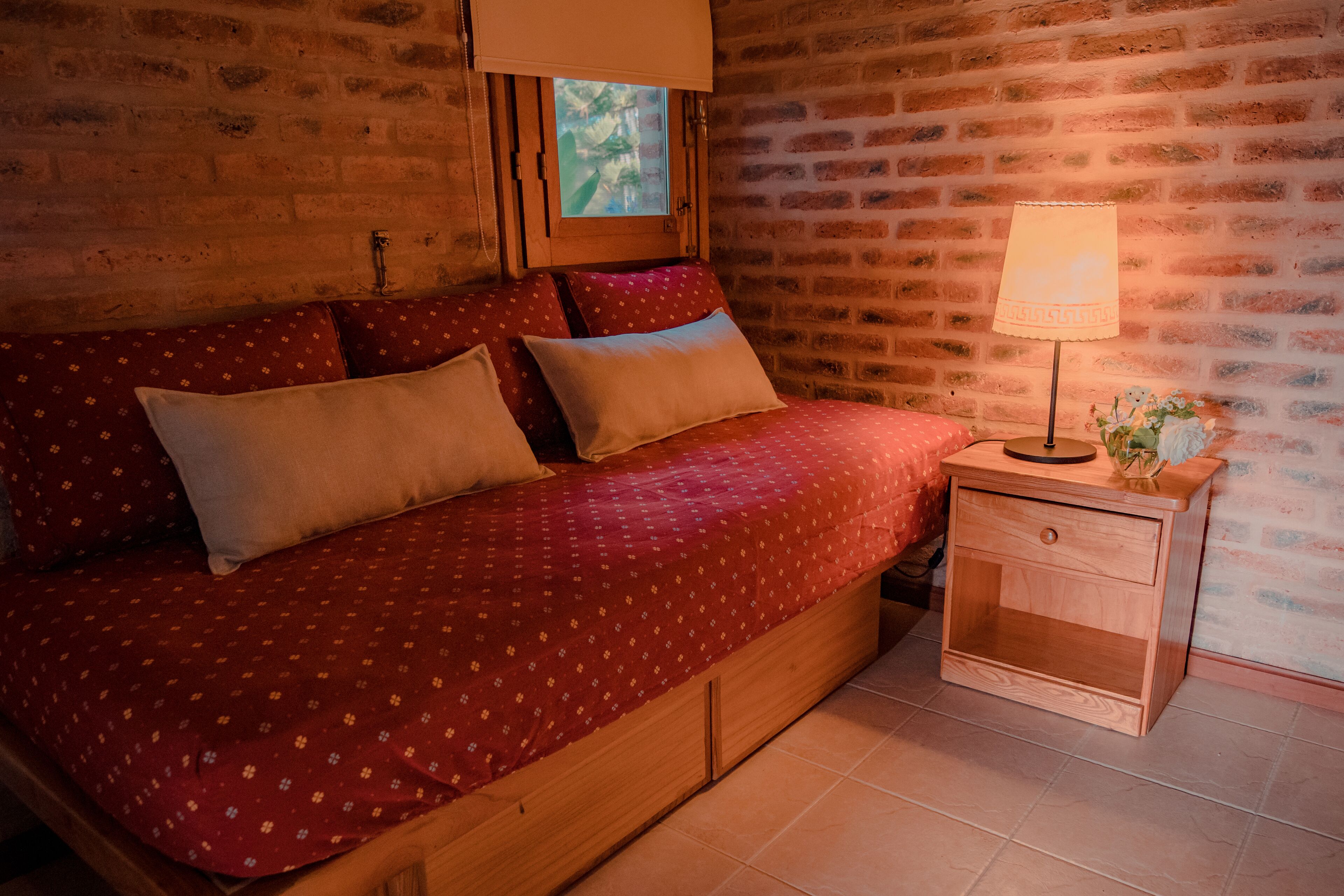 Una cama de madera con una colcha y almohadas estampadas en rojo, una mesita de noche con una lámpara, y una pared de ladrillo.