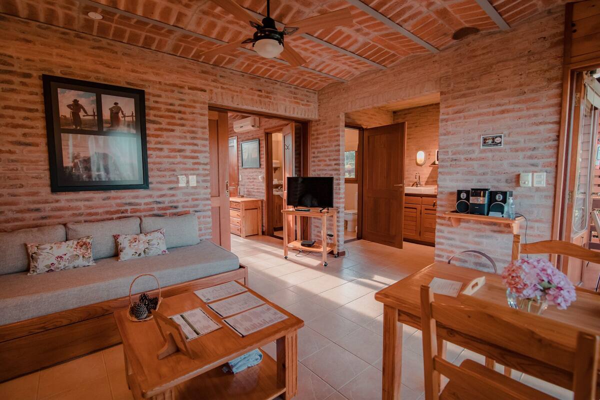 A cozy living room with a sofa, coffee table, and TV, connected to a kitchen area with wooden cabinets and a brick wall.