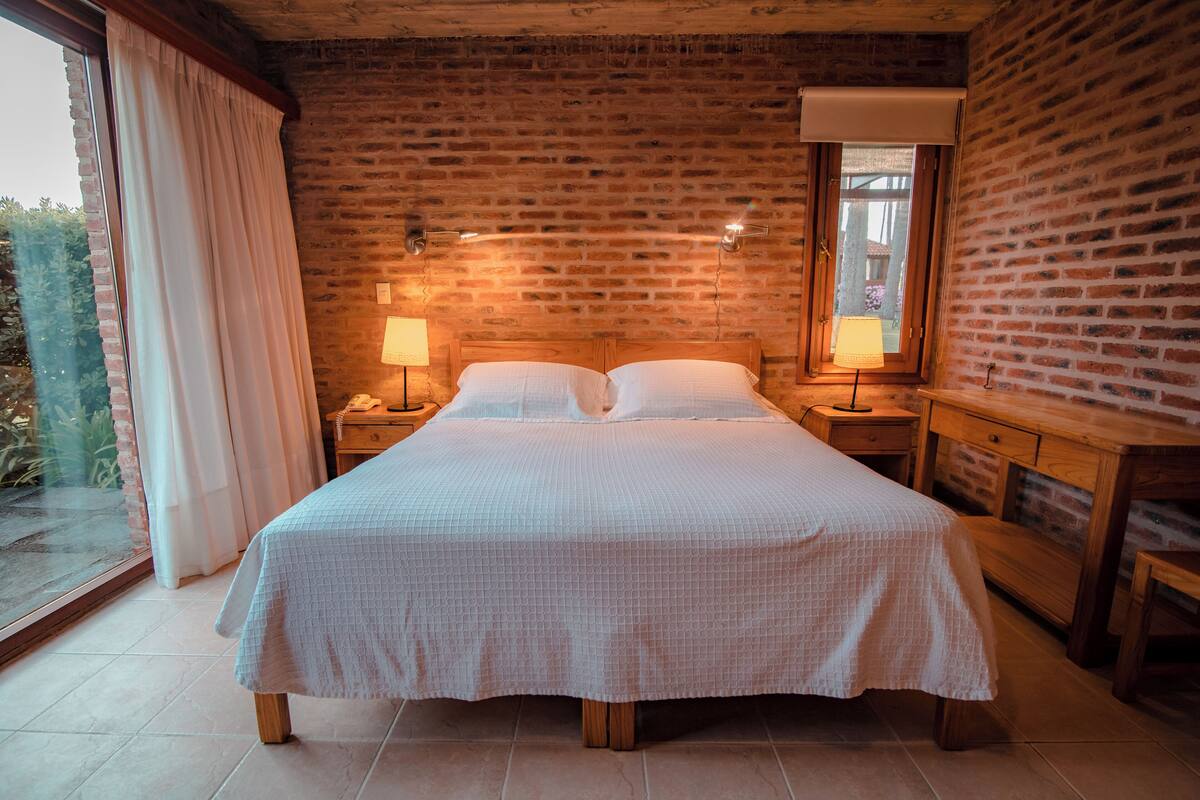 A bedroom with a large bed, wooden headboard, two bedside tables with lamps, a wooden dresser, and a window with curtains.