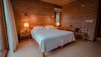 Un dormitorio con pared de ladrillo, cama con ropa blanca, dos mesitas de noche con lámparas, una silla de madera y una ventana con cortinas.
