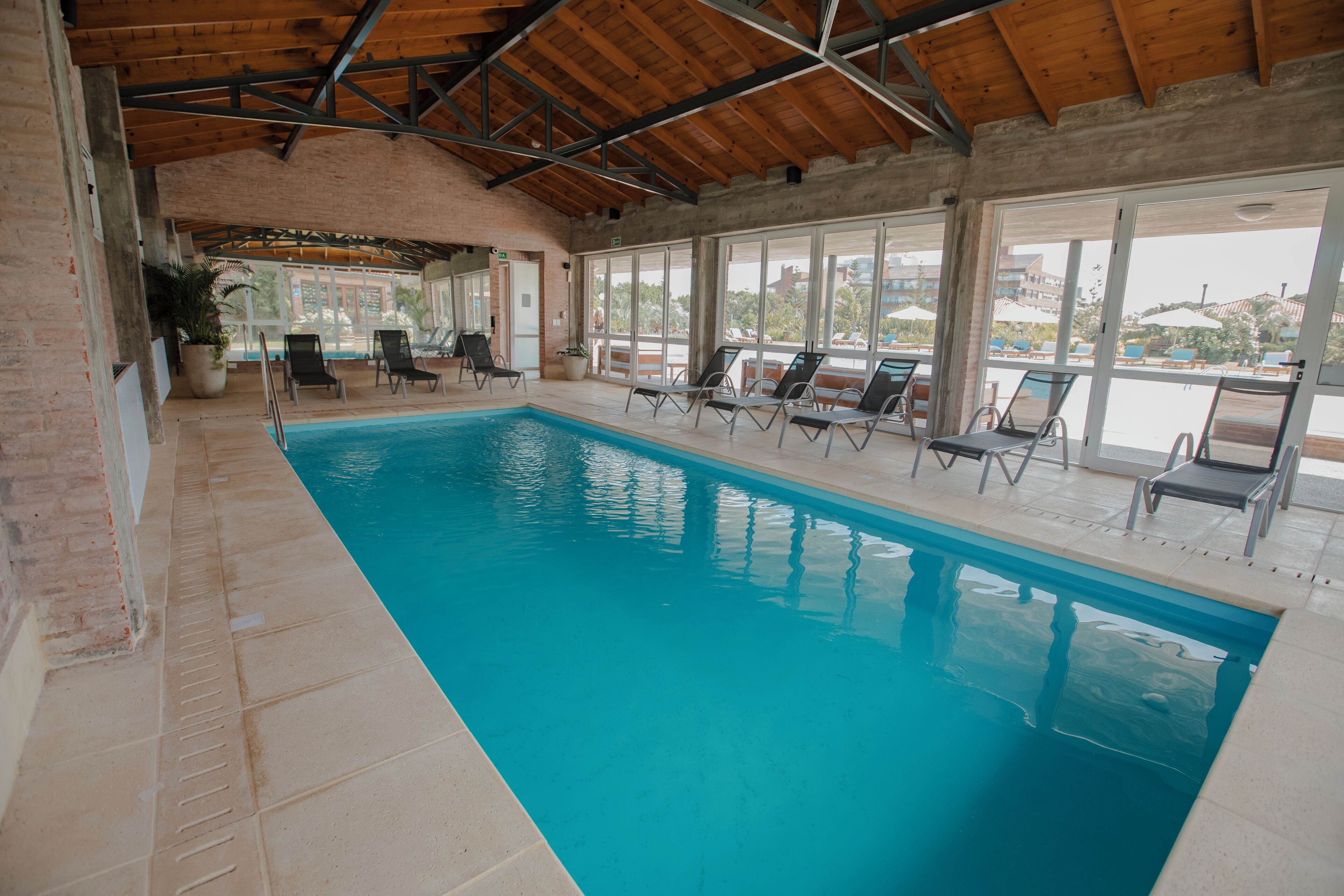 An indoor swimming pool with clear blue water, surrounded by lounge chairs and large windows offering views of the outside.