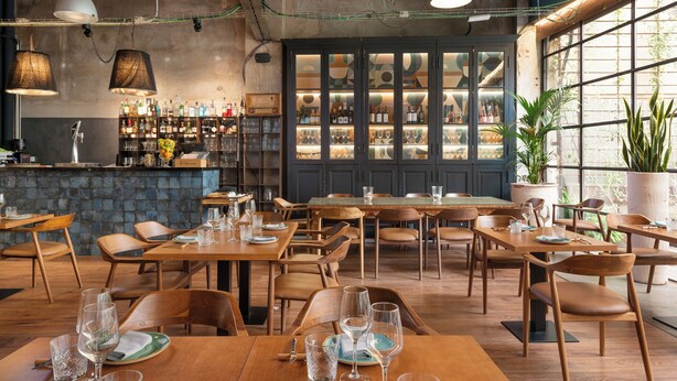Un restaurante con mesas y sillas de madera, una zona de bar con estanterías llenas de botellas y amplios ventanales que dejan entrar luz natural.