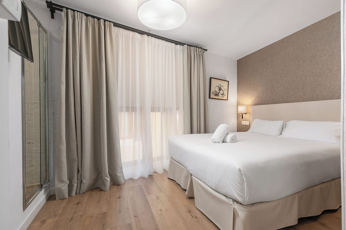 Habitación de hotel con una cama grande, una mesita de noche con lámpara, un cuadro enmarcado en la pared y una ventana con cortinas.