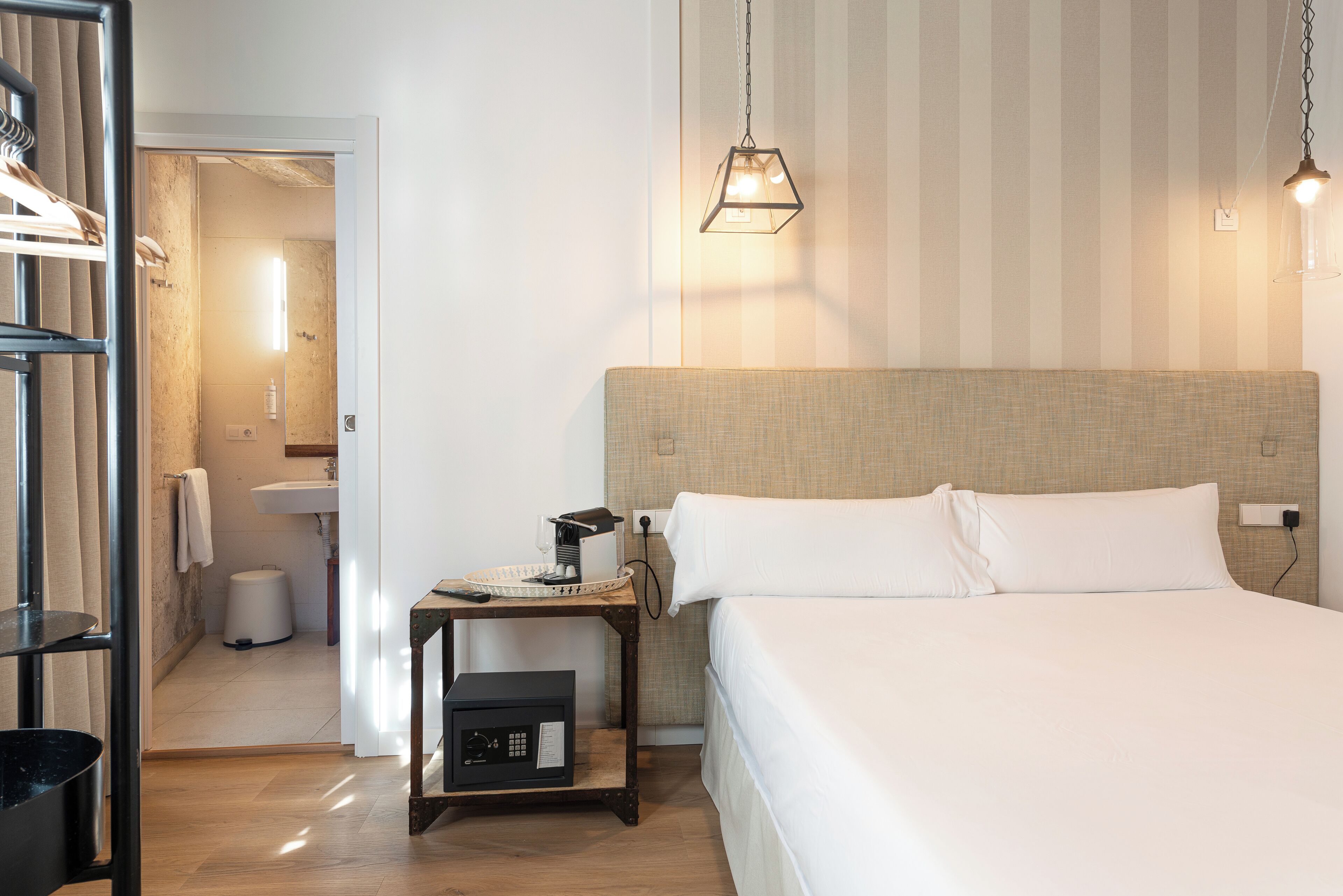 Habitación de hotel con una cama grande, una mesita de noche con lámpara, una pequeña mesa con teléfono y lámpara, un armario y un baño visible a través de una puerta abierta.
