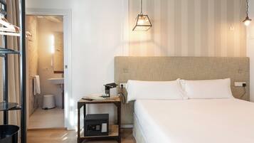 Habitación de hotel con una cama grande, una mesita de noche con lámpara, una pequeña mesa con teléfono y lámpara, un armario y un baño visible a través de una puerta abierta.