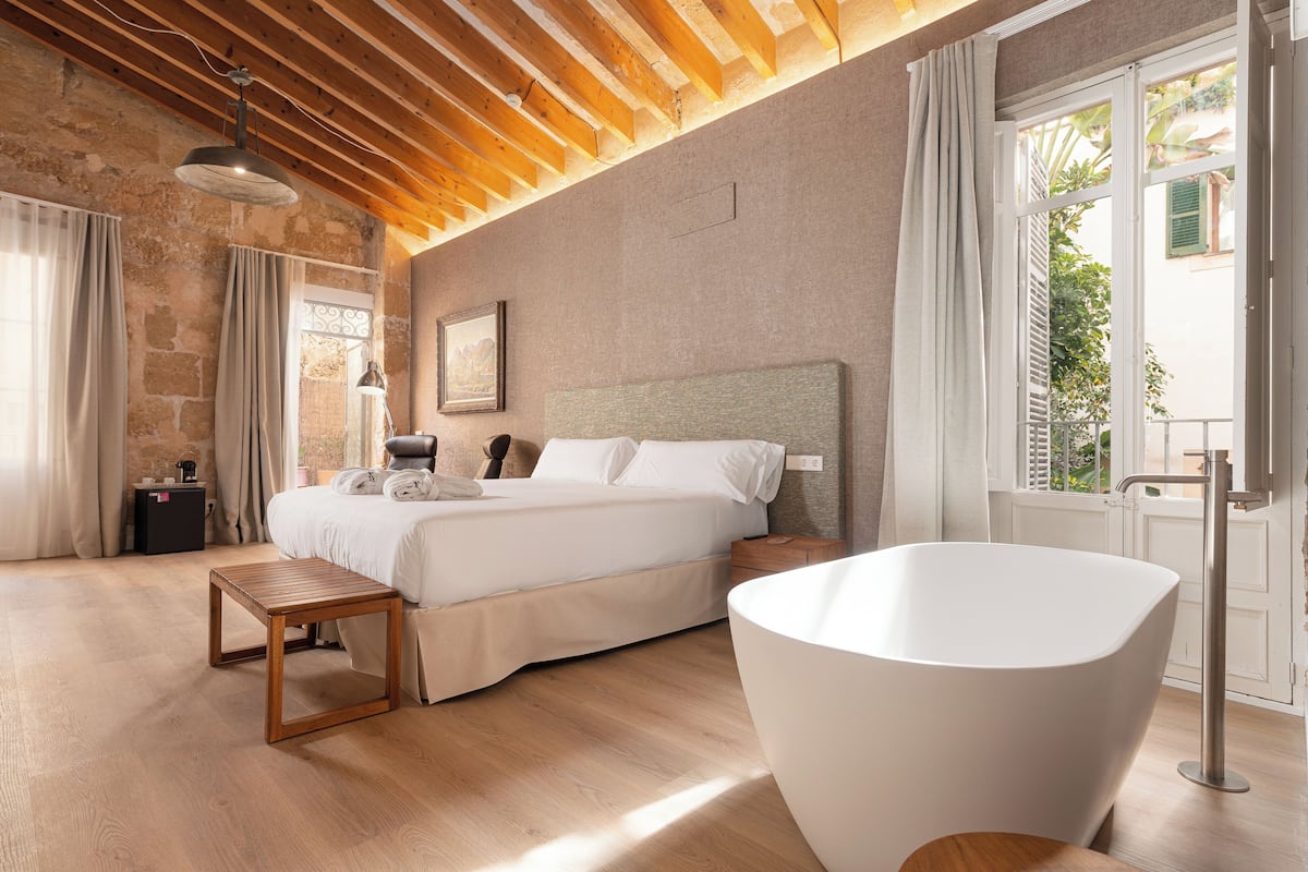Un dormitorio con una cama grande, una mesita de noche de madera, una bañera blanca y vistas a zonas verdes.