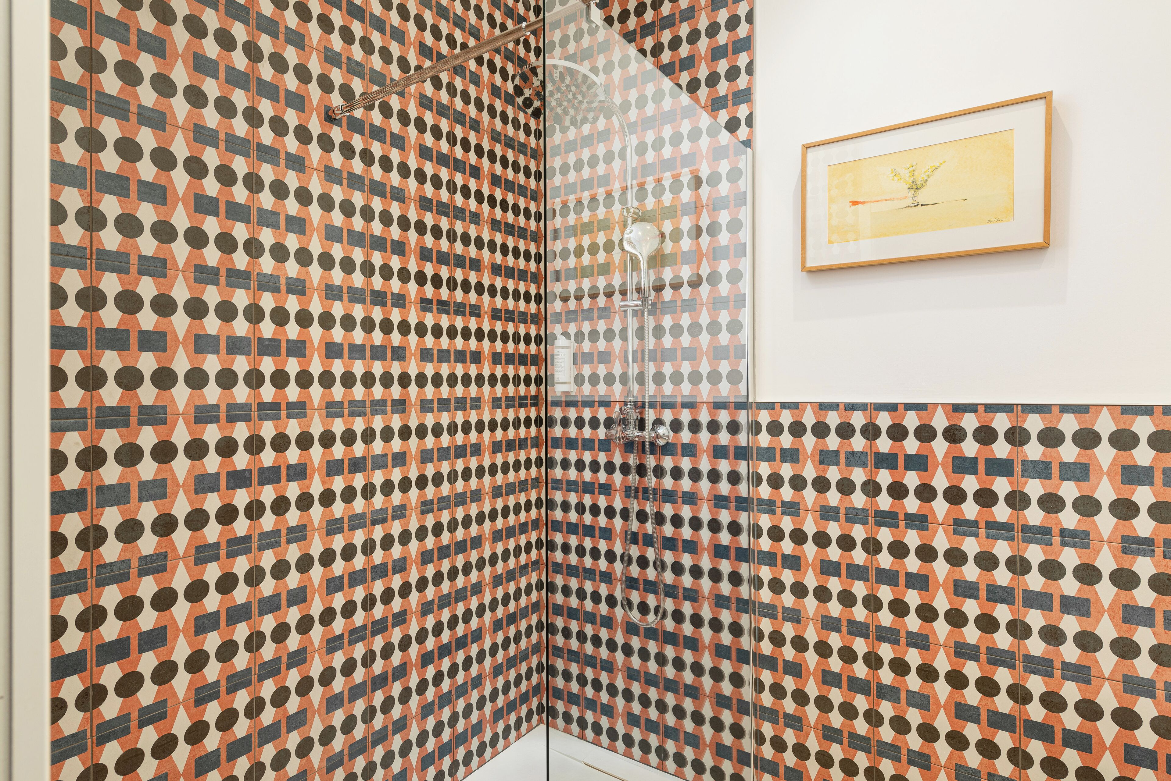 A bathroom with a geometric patterned tile design, a framed artwork on the wall, and a shower area.