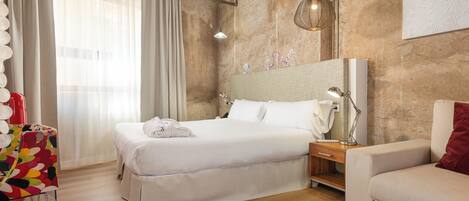 A hotel room with a bed, bedside table, lamp, chair, and a colorful chair.