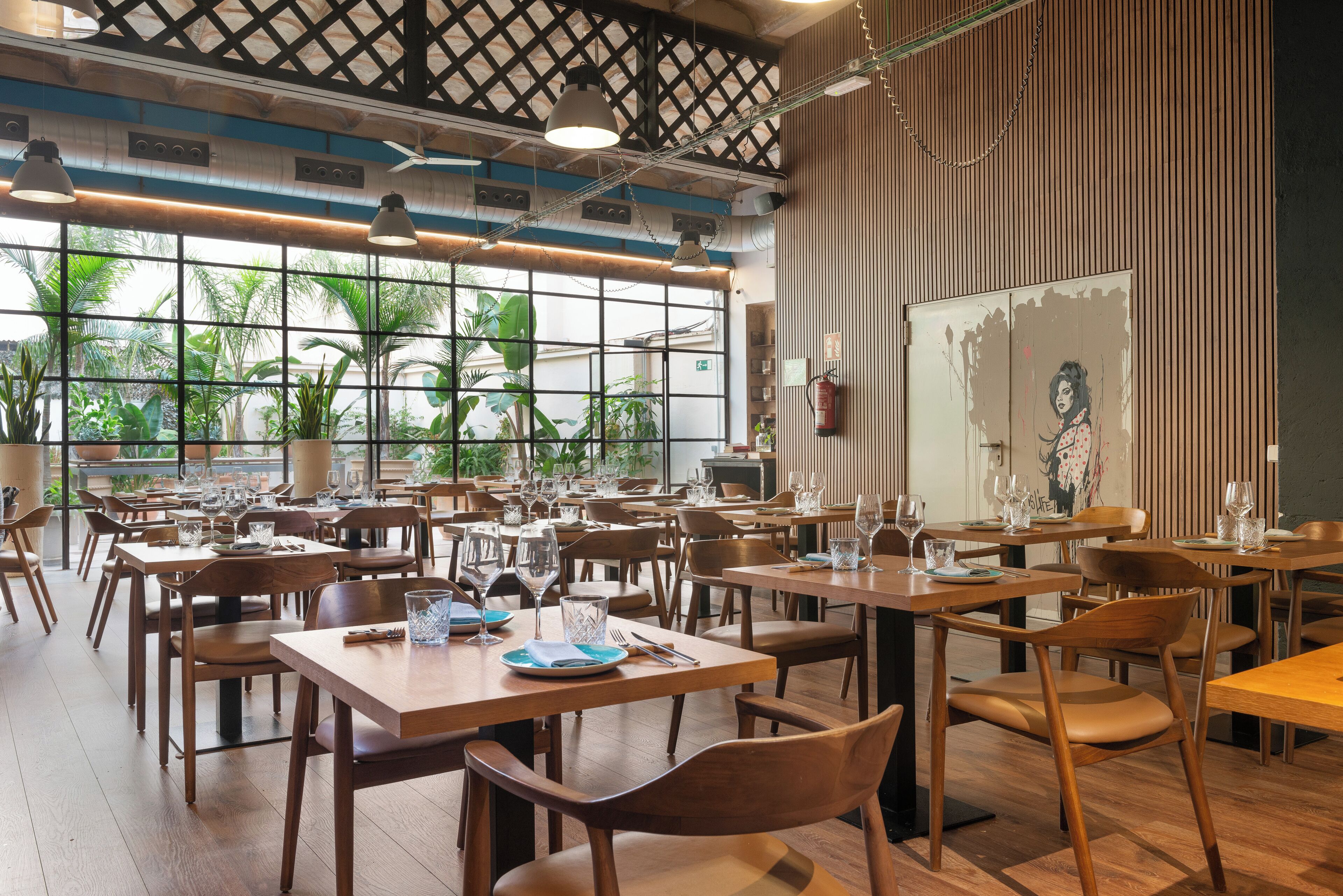 A restaurant with wooden tables and chairs, large windows with a view of greenery, and a modern ceiling design.