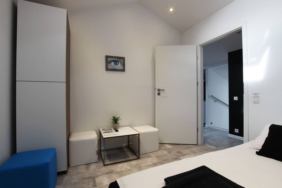 Una habitación de hotel moderna con una cama, una mesita con una planta, una silla azul y un armario blanco.