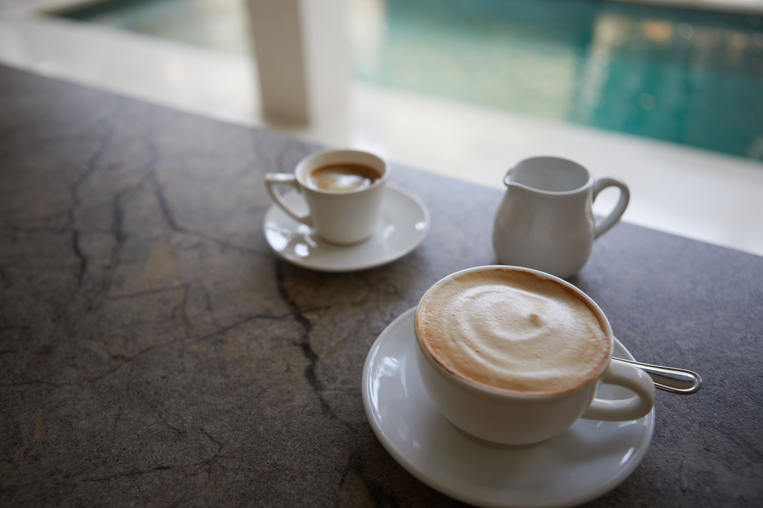 Ein Tisch mit zwei weißen Kaffeetassen und einer weißen Kanne auf einer Marmorplatte, nahe einem Fenster mit Blick auf einen Pool.