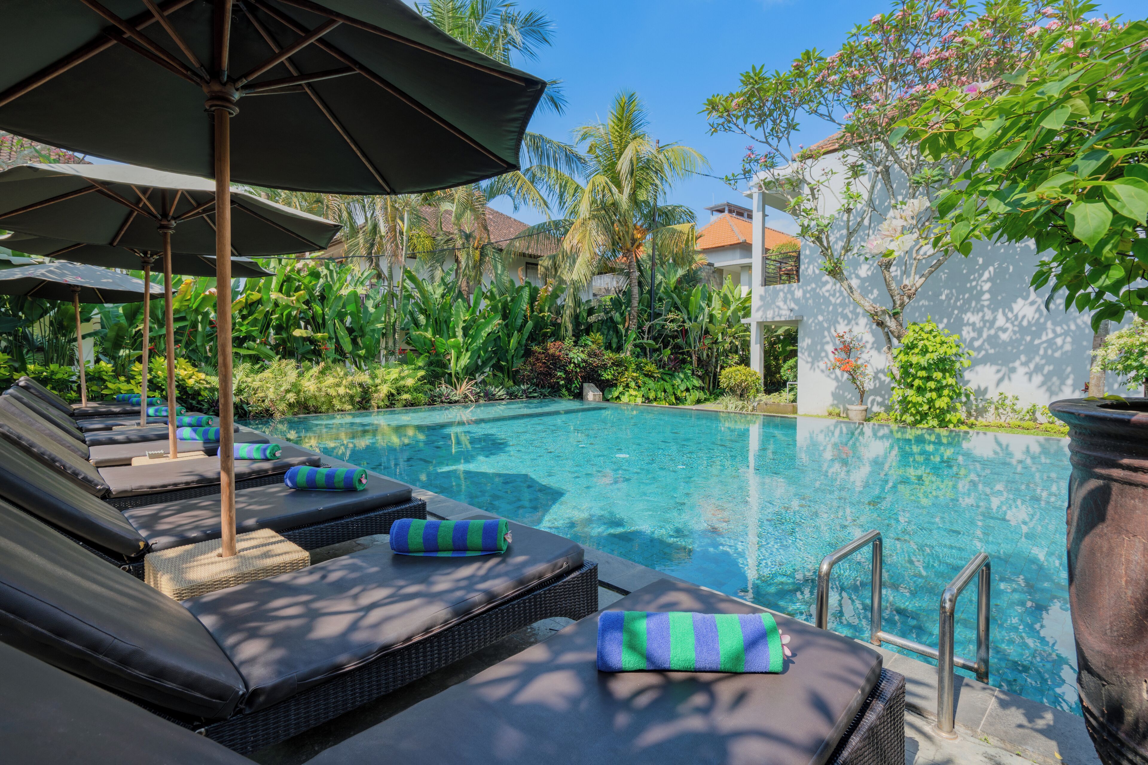 A poolside area with lounge chairs and umbrellas, surrounded by lush greenery and a clear blue pool.