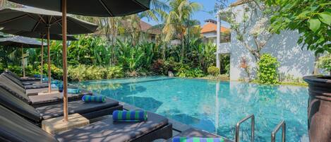 A poolside area with lounge chairs and umbrellas, surrounded by lush greenery and a clear blue pool.