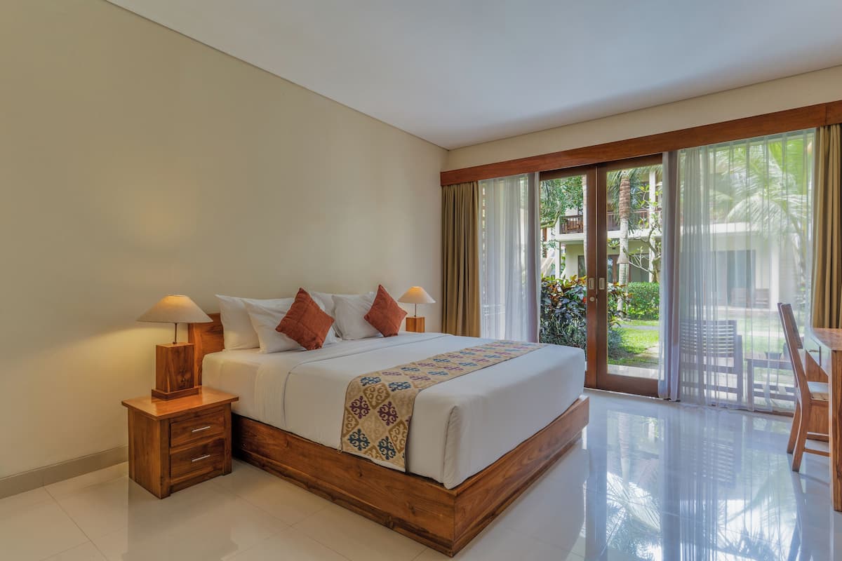 A hotel room with a large bed, wooden nightstands, a desk, and a view of a garden through the sliding glass door.