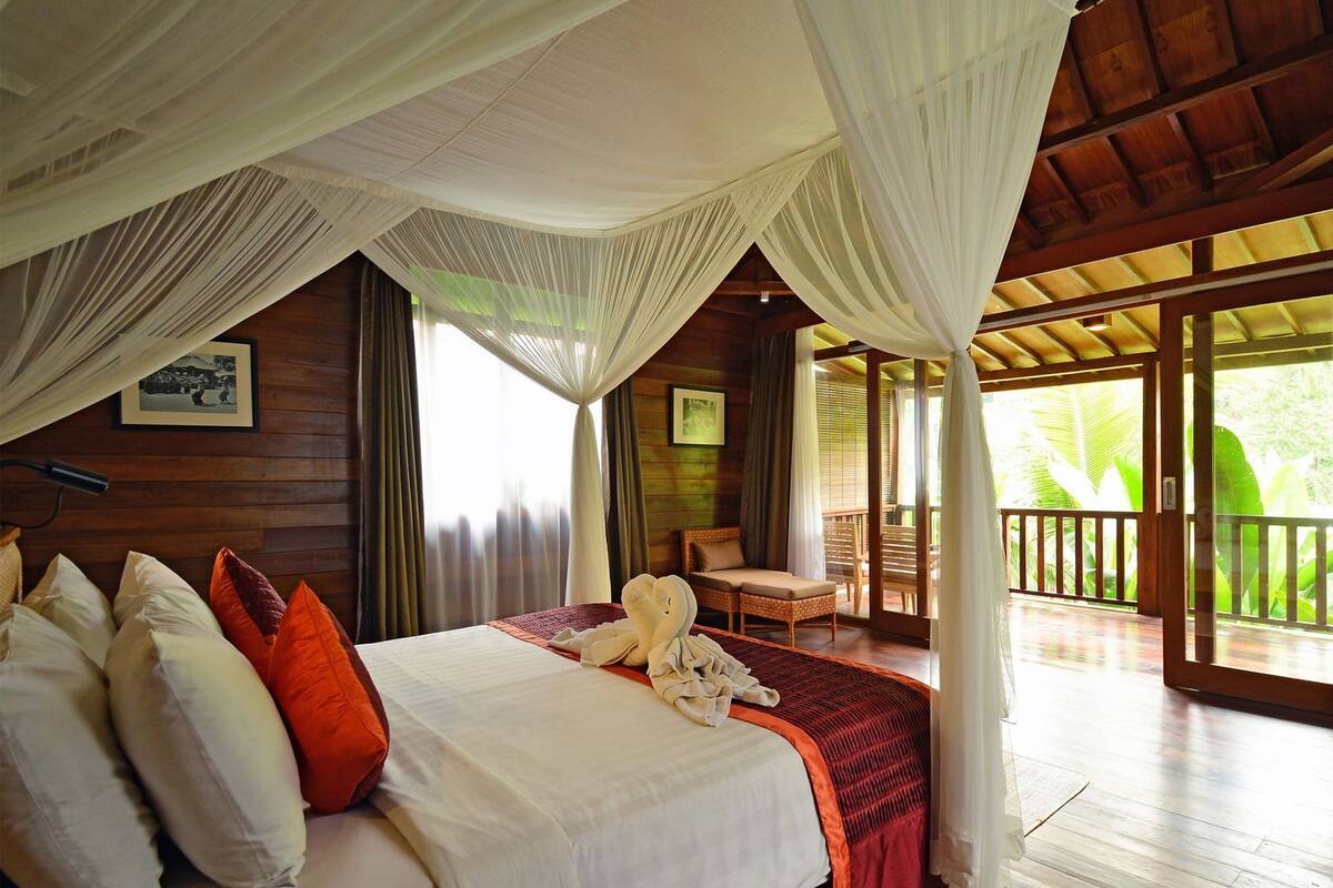 A bedroom with a bed, pillows, a bedside table, a chair, and a balcony with a view of greenery.